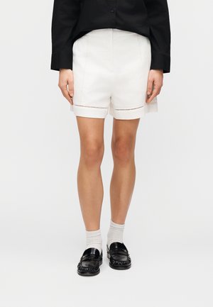 Białe szorty z przeszyciami i podwiniętymi mankietami, zestawione z czarnymi mokasynami i białymi skarpetkami, prezentujące czysty, minimalistyczny design.