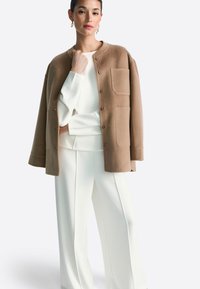 Brūns vilnas maisījuma žakete ar apaļu kakla izgriezumu, priekšējām pogām un divām kabatām, kombinēts ar baltu blūzi un platajiem baltiem biksēm.