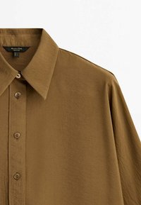 Chemise marron à boutons avec un col pointu, dotée d'une texture lisse, de six boutons et de manches légèrement surdimensionnées.