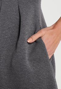 Grått tyg med en ribbad textur; visar en hand i en sidoficka, vilket framhäver plaggets fickdesign och materialdetalj.