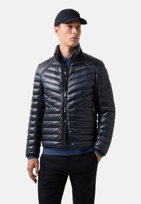 Námořnická puffer bunda s lesklým povrchem, prošívanou texturou, stojatým límcem, předním zipem a bočními kapsami. Nosič má na sobě modrou košili.