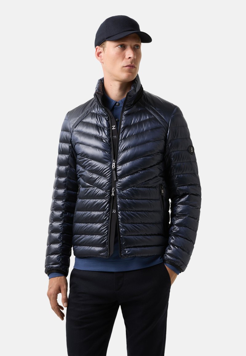 Námořnická puffer bunda s lesklým povrchem, prošívanou texturou, stojatým límcem, předním zipem a bočními kapsami. Nosi�č má na sobě modrou košili.