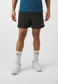 Pantalones cortos deportivos negros de tela ligera. Llevados con zapatillas blancas y calcetines con un logo de Puma negro. De corto largo, diseño casual.