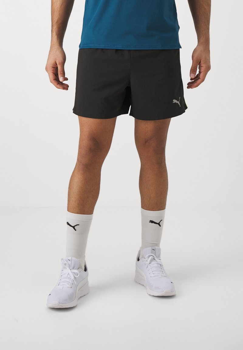 Pantalones cortos deportivos negros de tela ligera. Llevados con zapatillas blancas y calcetines con un logo de Puma negro. De corto largo, diseño casual.