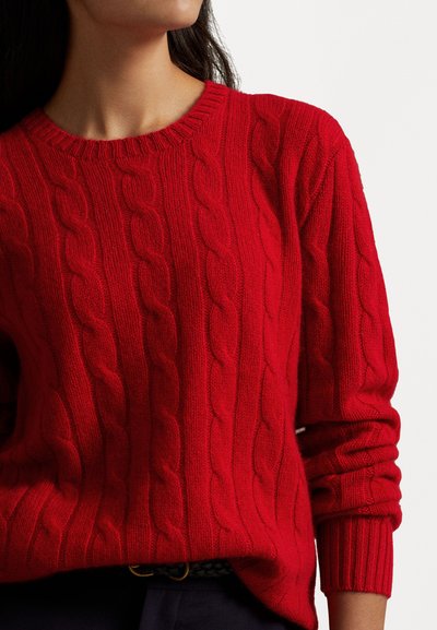 Suéter rojo de punto trenzado con cuello redondo y puños de canalé. Presenta un patrón texturizado y una tela suave, combinado con pantalones oscuros.