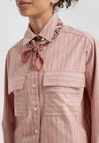 Chemise rose pâle à rayures verticales blanches, col noué avec une écharpe à motifs. Comprend deux poches poitrine avec fermetures à bouton.