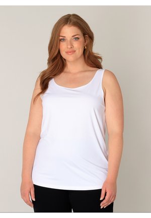 Vrouw met lang bruin haar draagt een mouwloos wit topje en zwarte broek, staand tegen een effen lichte achtergrond.