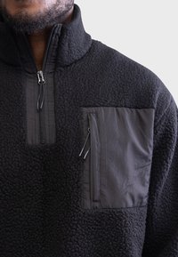 Pulover negru din fleece, cu guler cu jumătate de fermoar, țesătură texturată și o buzunar cu fermoar pe pieptul stâng dintr-un material contrastant.