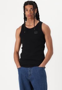 Črn top brez rokavov s okroglim izrezom in vezenim logotipom na prsih. Uporabljen s svetlo modrimi denim hlačami z ohlapnim krojem in žepi.