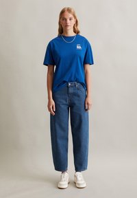 Blaues Baumwoll-T-Shirt mit kurzen Ärmeln und einem kleinen Grafikmotiv auf der Brust, kombiniert mit lässigen blauen Jeans und weißen Sneakers.