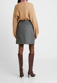 Nainen, joka pitää yllään beigeä cable knit -neuletta, harmaata villahametta ja ruskeita polven ylle ulottuvia korkokenkiä, seisoo selkä kameraan päin yksinkertaisella taustalla.