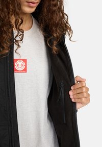 Person mit lockigem Haar, trägt ein graues Shirt mit rotem Logo "Wind-Wasser-Feuer-Erde" und zieht teilweise die schwarze Jacke auf, die eine Reißverschlusstasche im Inneren zeigt.