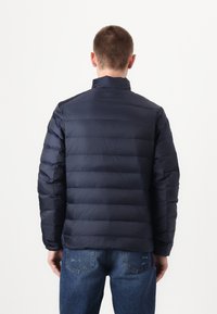 Jūrinės mėlynos spalvos pūkinė striukė su siuvinėtu dizainu, aukšta apykakle ir ilgomis rankovėmis. Pasižymi lygiu tekstūru ir minimalistinio dizaino elementais.