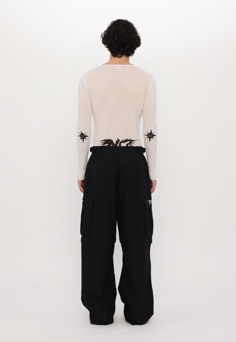 Top de manga larga en un beige claro con diseños de estrellas en las mangas; pantalones cargo negros con bolsillos laterales y un corte holgado. Vista trasera, fondo minimalista.