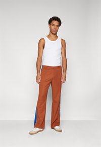 Bruine elastische broek met blauwe zijkanten, gecombineerd met een witte tanktop en witte sneakers. De stof lijkt zacht en textuur te hebben.