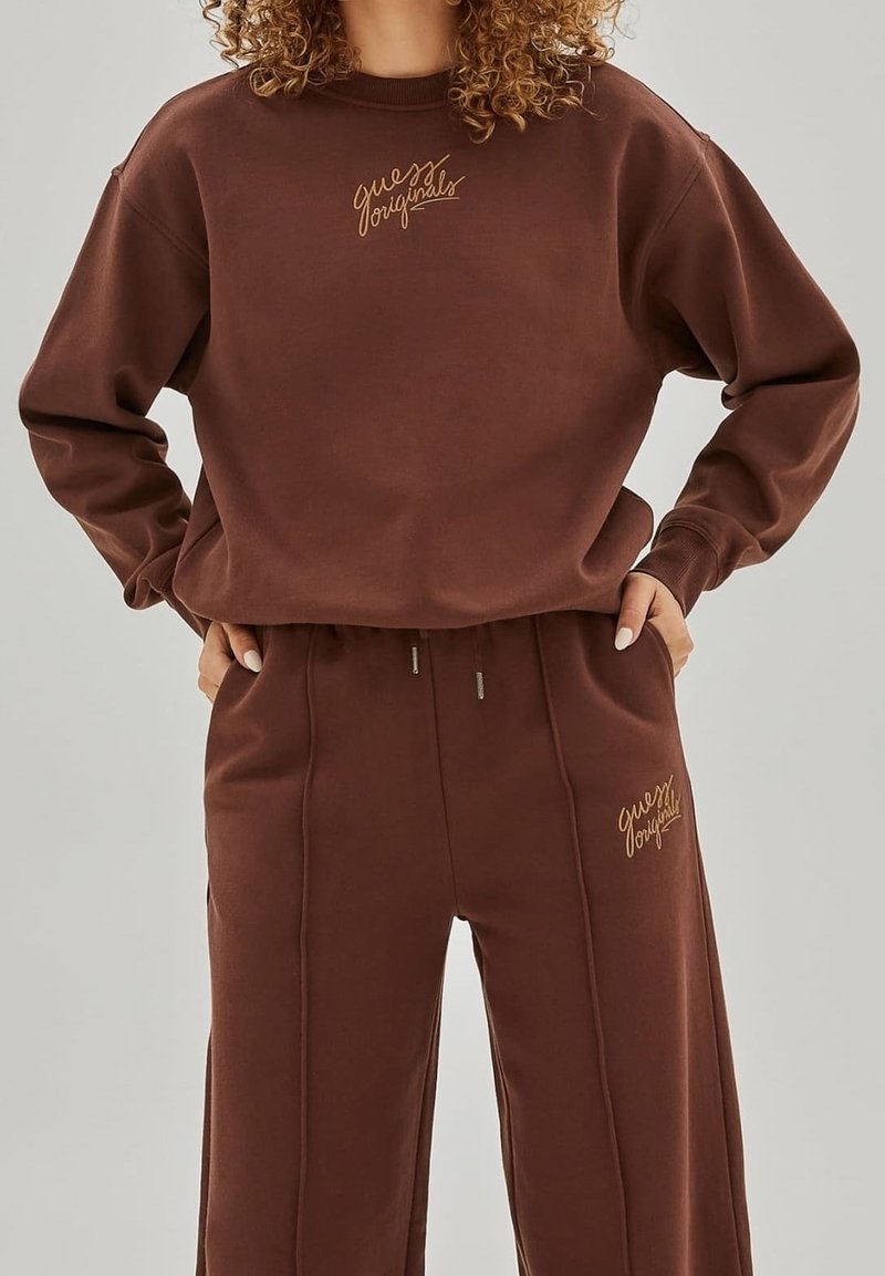 Sweatshirt marron et pantalon de jogging assorti en tissu doux, avec un logo brodé en or sur la poitrine et la cuisse.