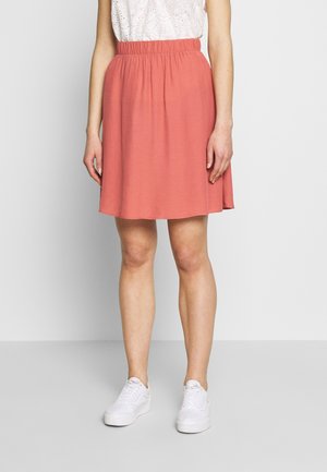 Femme portant une jupe corail longueur genou avec une taille élastique, un haut blanc à œillets et des baskets blanches, debout devant un fond uni.