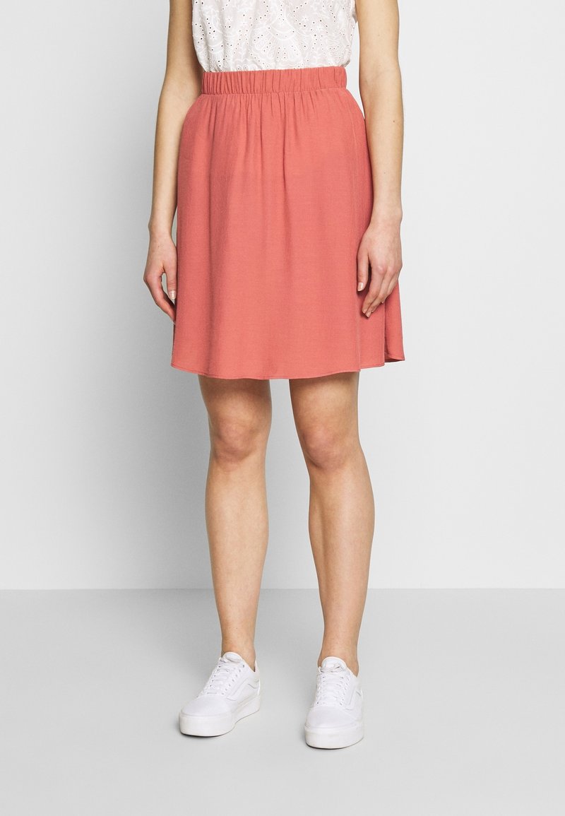 Femme portant une jupe corail longueur genou avec une taille élastique, un haut blanc à œillets et des baskets blanches, debout devant un fond uni.