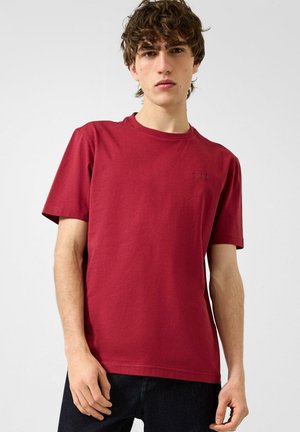 Junger Mann mit lockigen Haaren, der ein schlichtes rotes Kurzarm-T-Shirt und dunkle Hosen trägt und vor einem schlichten hellen Hintergrund steht.