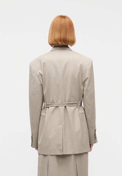 Άτομο με κοντά κόκκινα μαλλιά φορόντας μπεζ σακάκι με ριγέ λεπτομέρειες και ζώνη στο πίσω μέρος, από πίσω, με απλό φόντο.