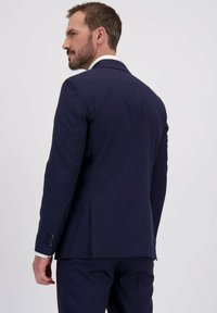 Man met een baard in een donkerblauw pak en een witte shirt staat iets gedraaid, kijkend naar zijn linkerzijde tegen een effen witte achtergrond.