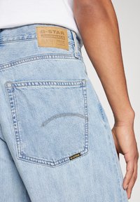 Svijetloplave traperice s bež kožnom oznakom s natpisom "G-STAR ORIGINALS RAW DENIM." Imaju vidljive šavove i dizajn stražnjeg džepa.