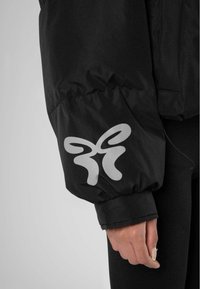 Zwarte gewatteerde jas met een zilveren vlinderlogo op de mouw. De stof lijkt glad met een licht glanzende textuur.
