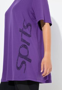 Fioletowy sportowy top z krótkim rękawem z czarnym napisem w wzór zebry po boku. Wykonany z gładkiego, elastycznego materiału o luźnym kroju.