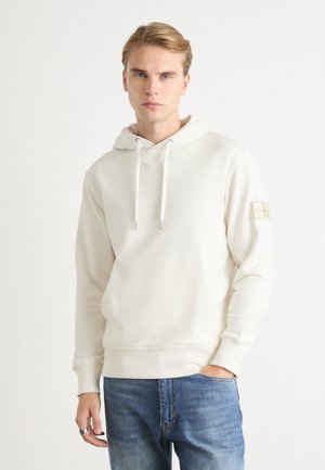 Crème hoodie met een voorzak, trekkoordkap, geribbelde manchetten en een klein logopatch op de linkermouw. Draagt met blauwe jeans.