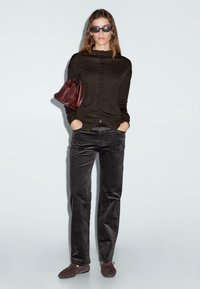 Donna con un top nero con bottoni, pantaloni di velluto neri, scarpe scure, occhiali da sole e che tiene in mano una borsa a tracolla rossa scura, con le mani nelle tasche.