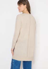 Femme portant un long cardigan beige en maille avec des détails côtelés verticaux dans le dos et sur les manches, assorti à un jean bleu, sur fond blanc.
