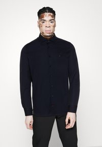 Mann mit Vitiligo, der ein schwarzes Hemd mit Knöpfen und schwarze Hosen trägt, steht vor einem schlichten hellen Hintergrund.