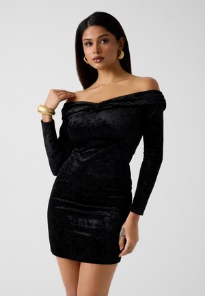 Vrouw die een zwarte fluwelen off-shoulder jurk draagt, met gouden oorringen en een gouden armband, poserend tegen een neutrale achtergrond.