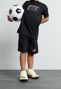 Camiseta negra de Nike con logo blanco, pantalones cortos negros, botines de fútbol amarillos con detalles negros, sosteniendo un balón de fútbol blanco y negro.