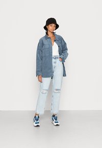 Camisa de ganga em azul claro, com grandes bolsos frontais. Usada por cima de uma blusa branca, com jeans em azul claro de cintura alta e desgastados, e calçado desportivo.