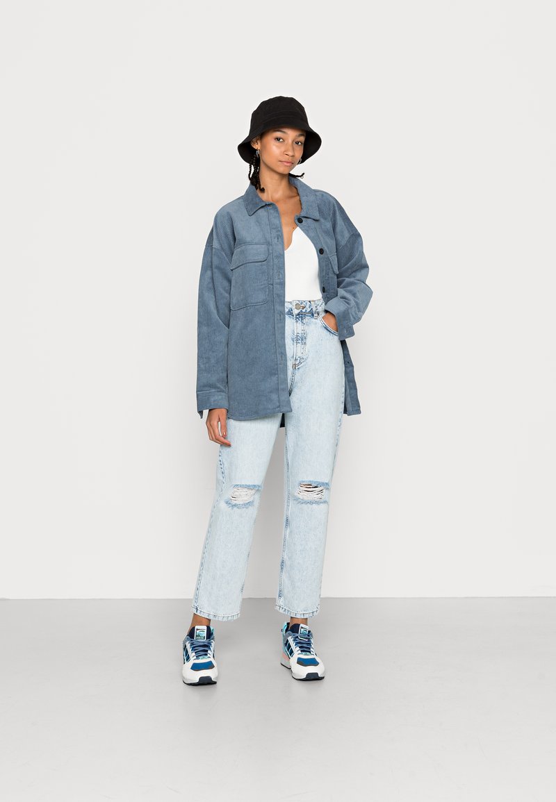 Hellblaues Denimhemd mit großen Fronttaschen. Trägt man über einem weißen Oberteil mit hochgeschnittenen, zerrissenen hellblauen Jeans und sportlichem Schuhwerk.