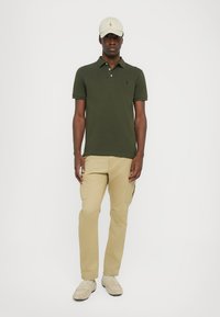 Maslinastozelena polo majica, bež cargo hlače, bež kapa i svijetle cipele, prikazujući ležeran, opušten outfit s klasičnim krojem i teksturama.