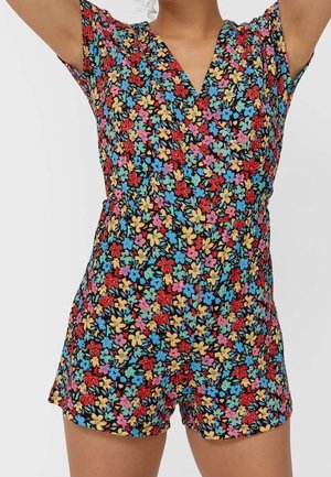 Frau trägt einen farbenfrohen, kurzärmeligen Jumpsuit mit Blumenmuster in Rot, Blau, Gelb und Pink auf schwarzem Hintergrund.