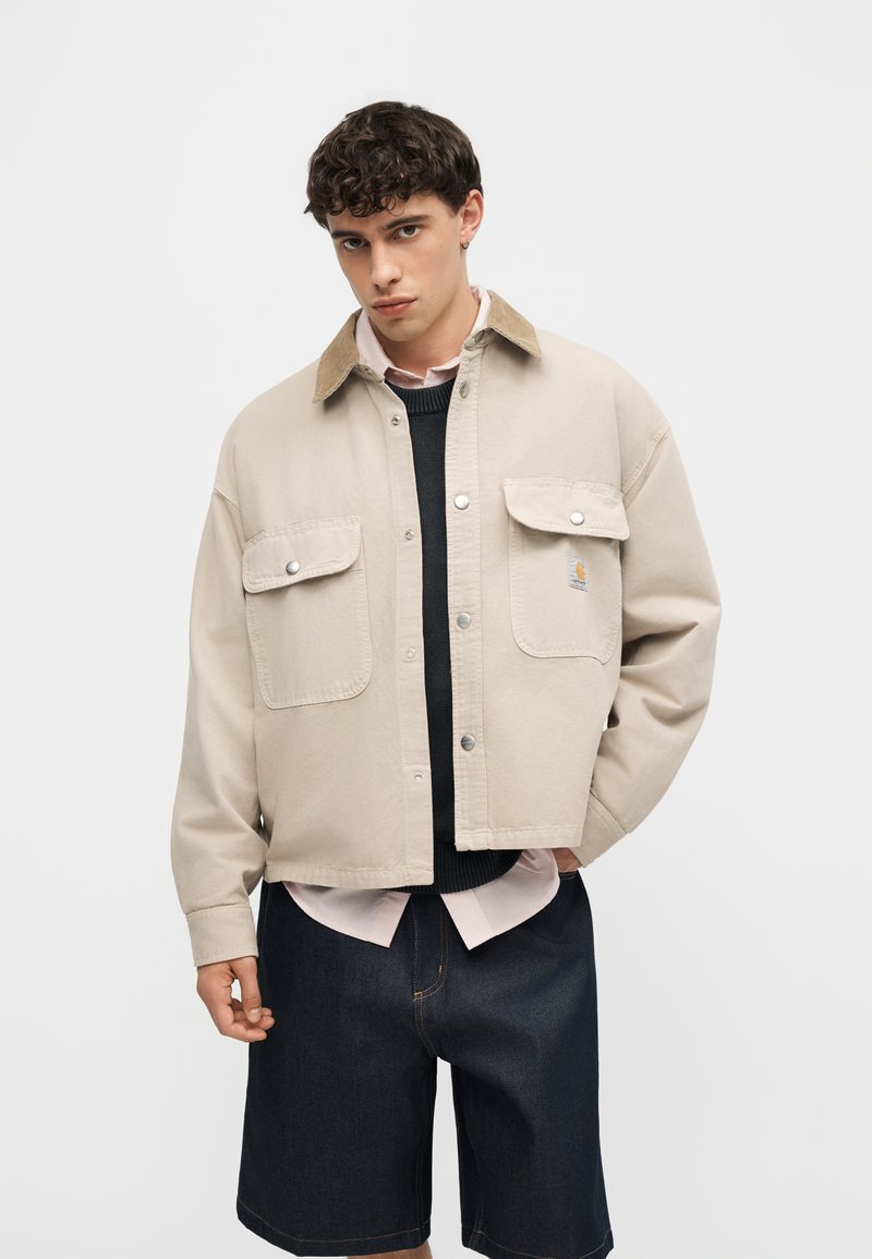 Joven con pelo rizado que lleva una chaqueta beige sobre un suéter negro y una camisa rosa, combinado con pantalones cortos de mezclilla oscuros hasta la rodilla.