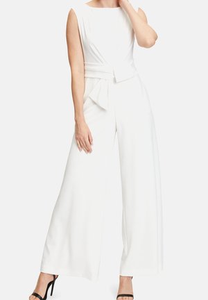 Kobieta ubrana w białe, bezrękawnikowe kombinezon z szerokimi nogawkami i warstwową warstwą w talii oraz czarne sandały na wysokim obcasie na jasnym tle.