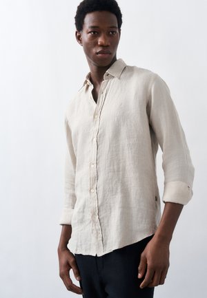 Junger Mann trägt ein beiges Hemd mit langen Ärmeln und aufgerollten Ärmeln, steht vor einem schlichten weißen Hintergrund.