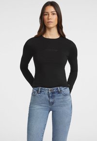 Crni dugih rukava uski top s kružnim izrezom, s diskretnim 'Guess Jeans' brandingom na prsima. Uparen s svijetloplavim trapericama.