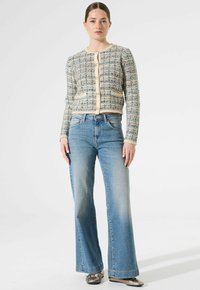 Una mujer está de pie vistiendo un cárdigan abotonado de patrón azul y beige, jeans de pierna ancha de color azul claro y zapatos metálicos de punta, contra un fondo blanco.