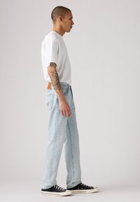Jeans dritti blu chiaro con una toppa in pelle, abbinati a una t-shirt bianca e sneakers nere basse. Tessuto testurizzato, vestibilità casual.