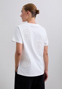 Femme avec un chignon portant un T-shirt blanc orné de trois ronds en crochet dans le dos, associé à un pantalon noir sur fond gris.