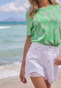 Mujer vestida con una camiseta de patrón ondulado verde y rosa y pantalones cortos de mezclilla blancos desgastados, de pie junto a la playa con las olas del océano de fondo.