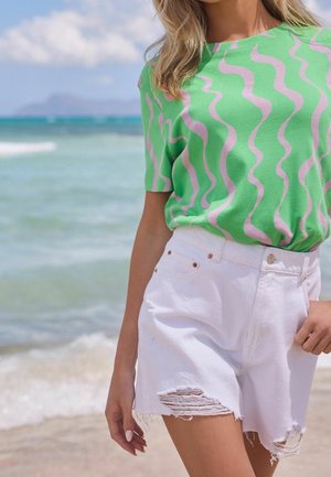 Femme portant une chemise à motifs en vagues verts et roses et un short en denim blanc déchiré, se tenant près de la plage avec des vagues de l'océan en arrière-plan.