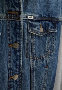 Zbliżenie na kieszeń niebieskiej kurtki jeansowej z metalowym guzikiem i małą białą metką z napisem „GUESS Originals”.