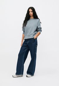 Sudadera gris de corte holgado, combinada con jeans de pierna ancha en azul oscuro. Incluye mangas con puños y textura de mezclilla. El calzado son zapatillas blancas.