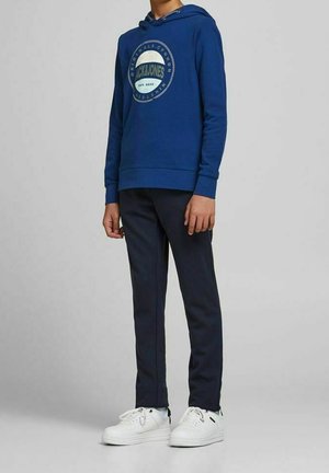 Niebieska bawełniana bluza z kapturem z okrągłym graficznym logo, zestawiona z ciemnonawigacyjnymi spodniami i białymi sneakersami. Prosty design, standardowy krój.